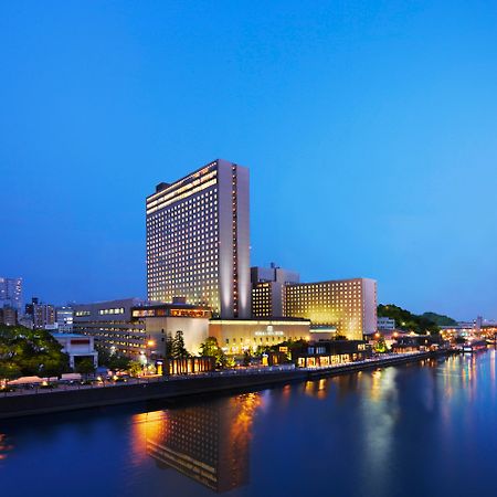 Rihga Royal Hotel Osaka, Vignette Collection By Ihg Extérieur photo The photo shows a large hotel building situated near a body of water, likely a river. The scene is illuminated by soft evening lighting, giving the environment a tranquil, serene feel. In the background, there are various buildings and structures, su