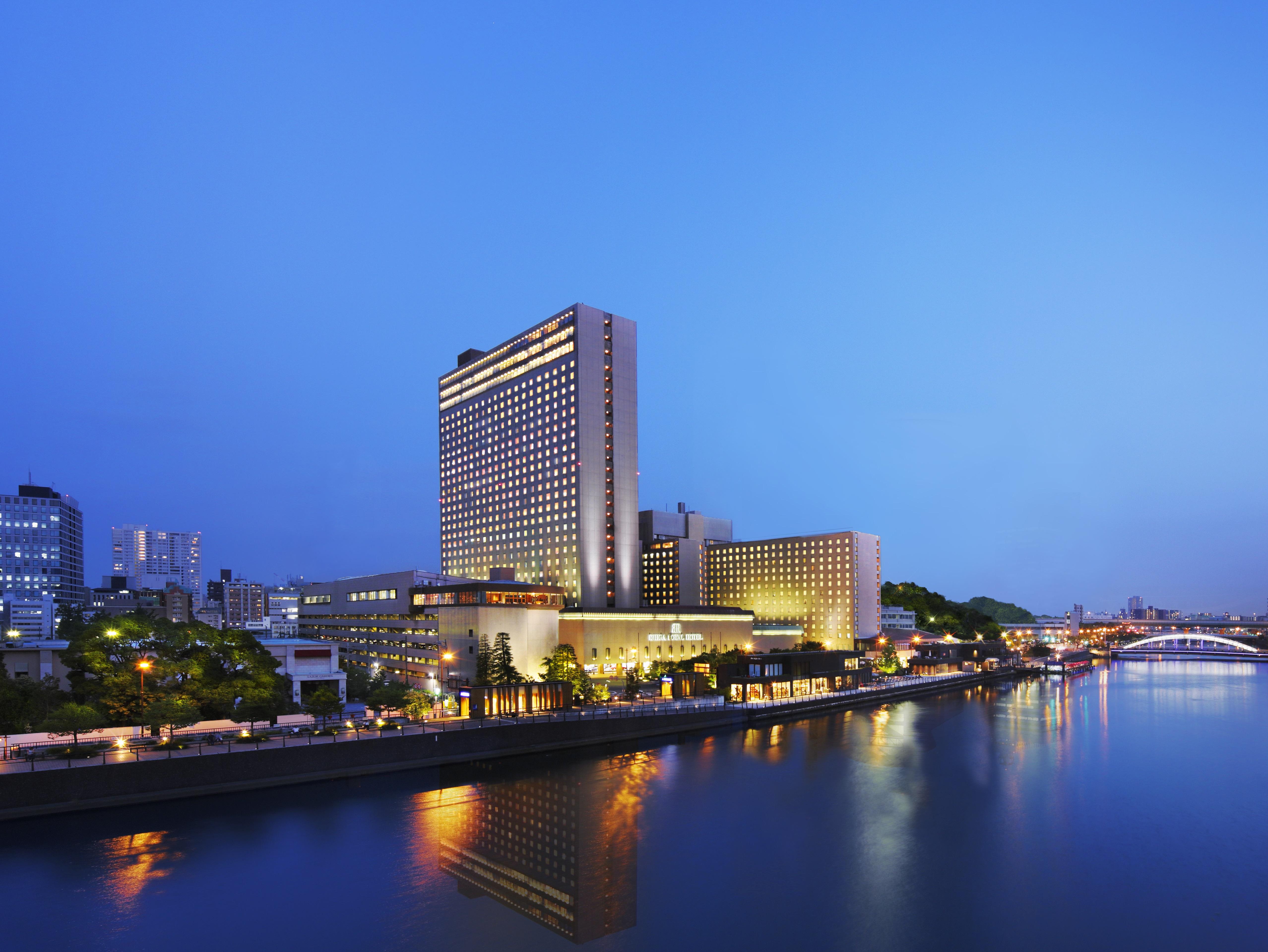 Rihga Royal Hotel Osaka, Vignette Collection By Ihg Extérieur photo The photo shows a large hotel building situated near a body of water, likely a river. The scene is illuminated by soft evening lighting, giving the environment a tranquil, serene feel. In the background, there are various buildings and structures, su
