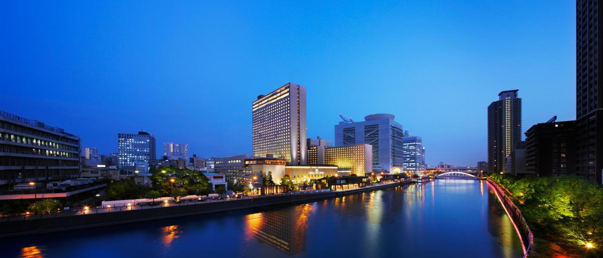 Rihga Royal Hotel Osaka, Vignette Collection By Ihg Extérieur photo The photo shows a cityscape at dusk, featuring a modern hotel and several tall buildings along a river. The structures are illuminated with lights, reflecting beautifully on the water. The atmosphere has a serene and urban feel, with a clear blue sky